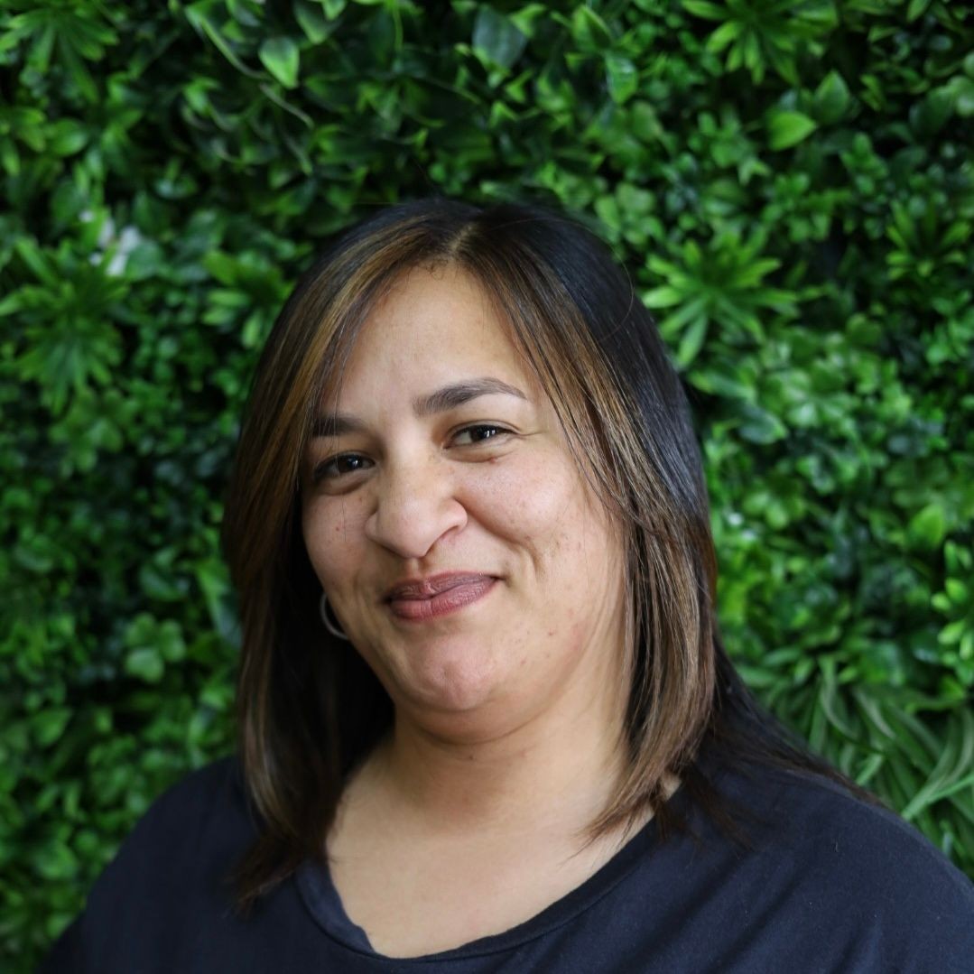 Smiling person with shoulder-length hair in front of a lush green plant wall.