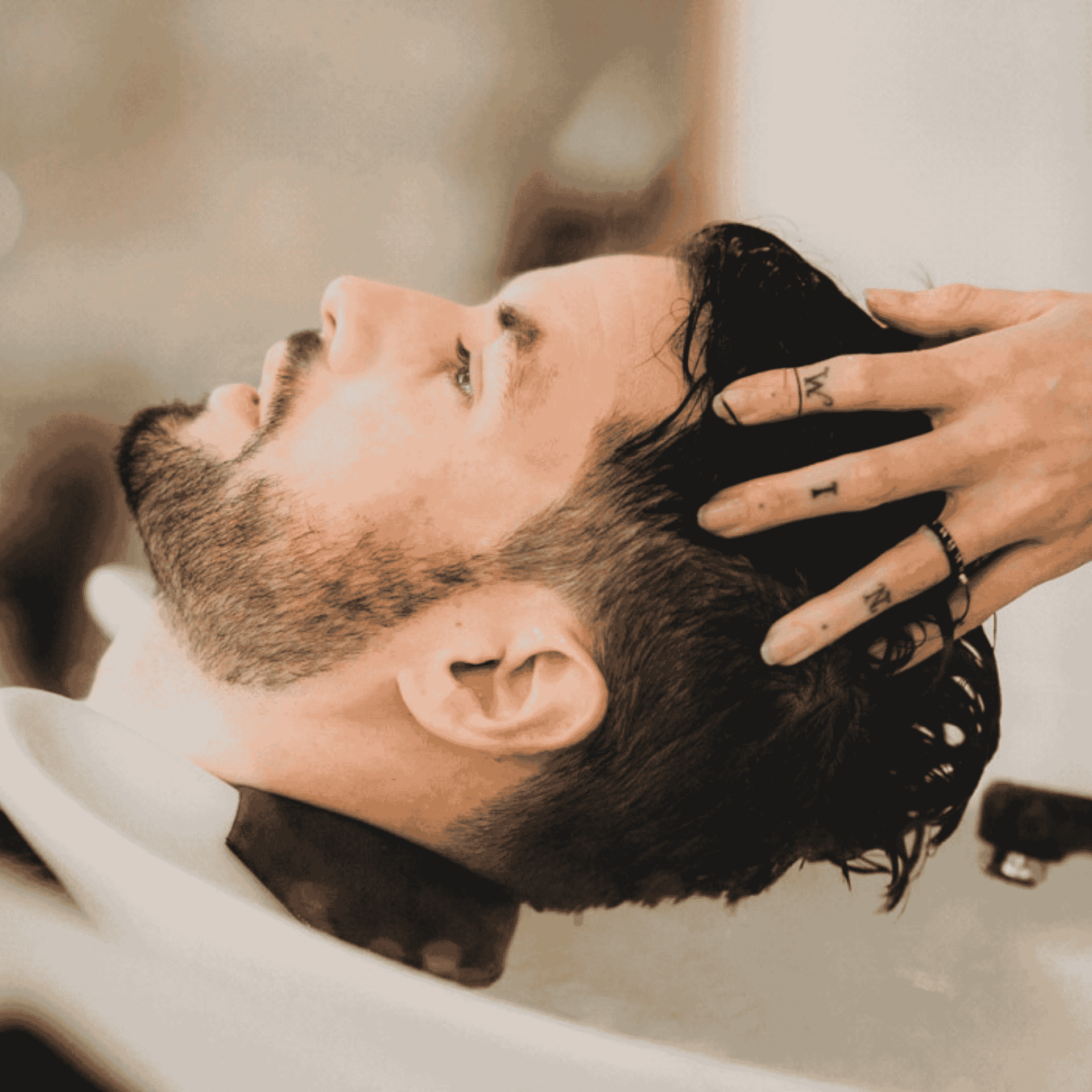 Man with beard getting a hair wash in a salon sink while someone massages his scalp.