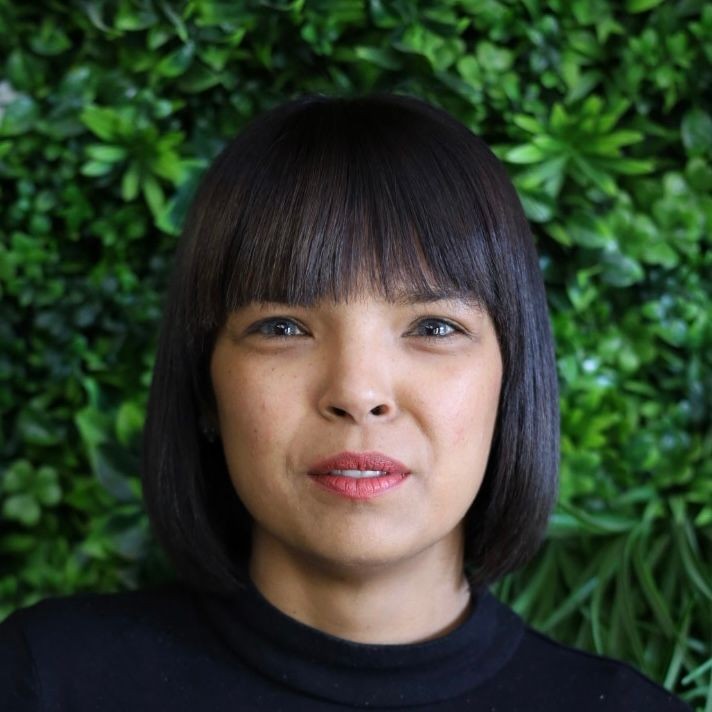 Person with dark hair and bangs in front of a green leafy background.