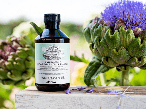 Bottle of detoxifying scrub shampoo on wooden surface with purple flower and artichoke in background.