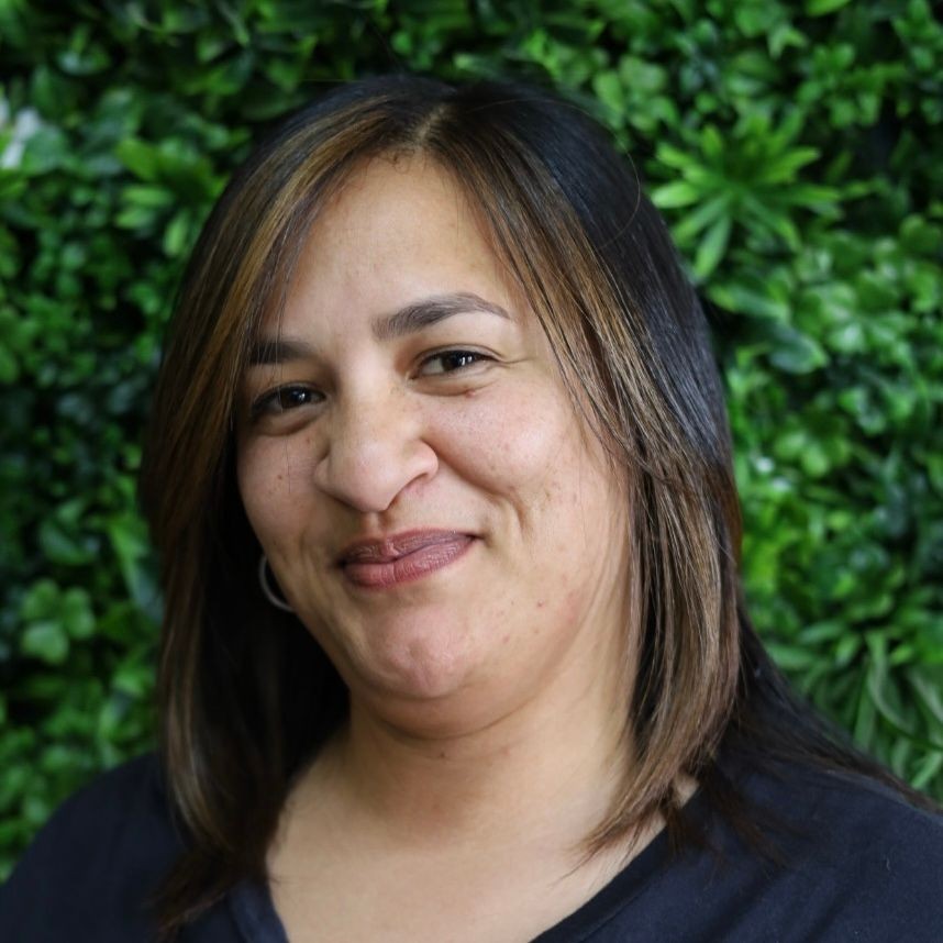Person with shoulder-length hair standing in front of a wall covered in green foliage.