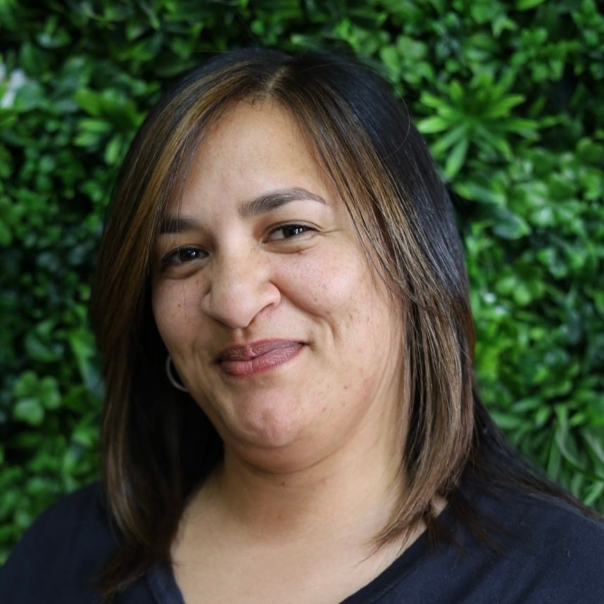 Person with shoulder-length hair standing in front of a wall covered in green foliage.